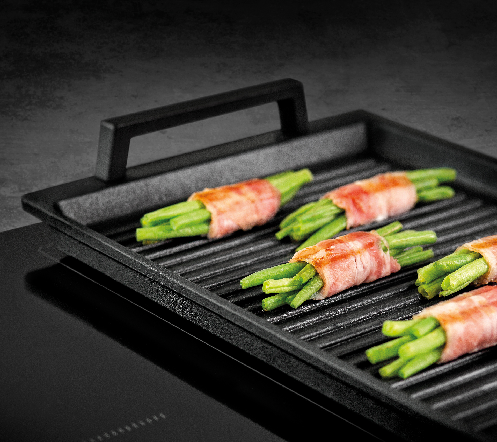 Eine schwarze Grillplatte in Nahaufnahme auf einer Kochfläche mit grünen Bohnen im Speckmantel